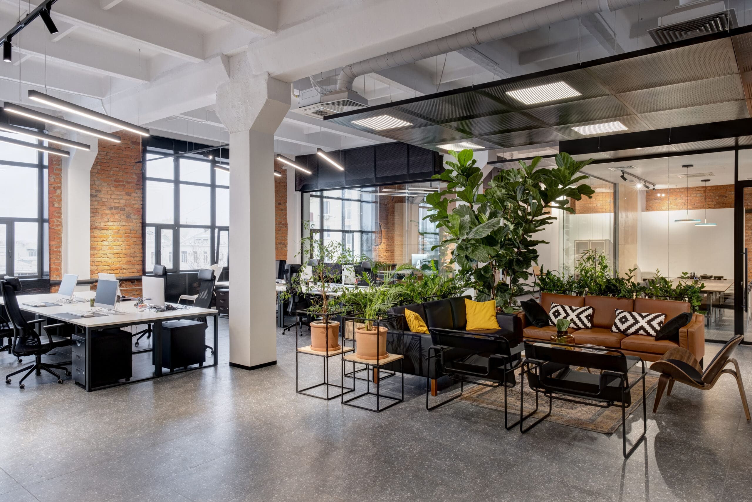 Modern office space with large windows, exposed brick walls, and a mix of workstations and a lounge area. The lounge features a brown leather sofa, chairs, and green plants. Hanging lights illuminate the clean, open environment.