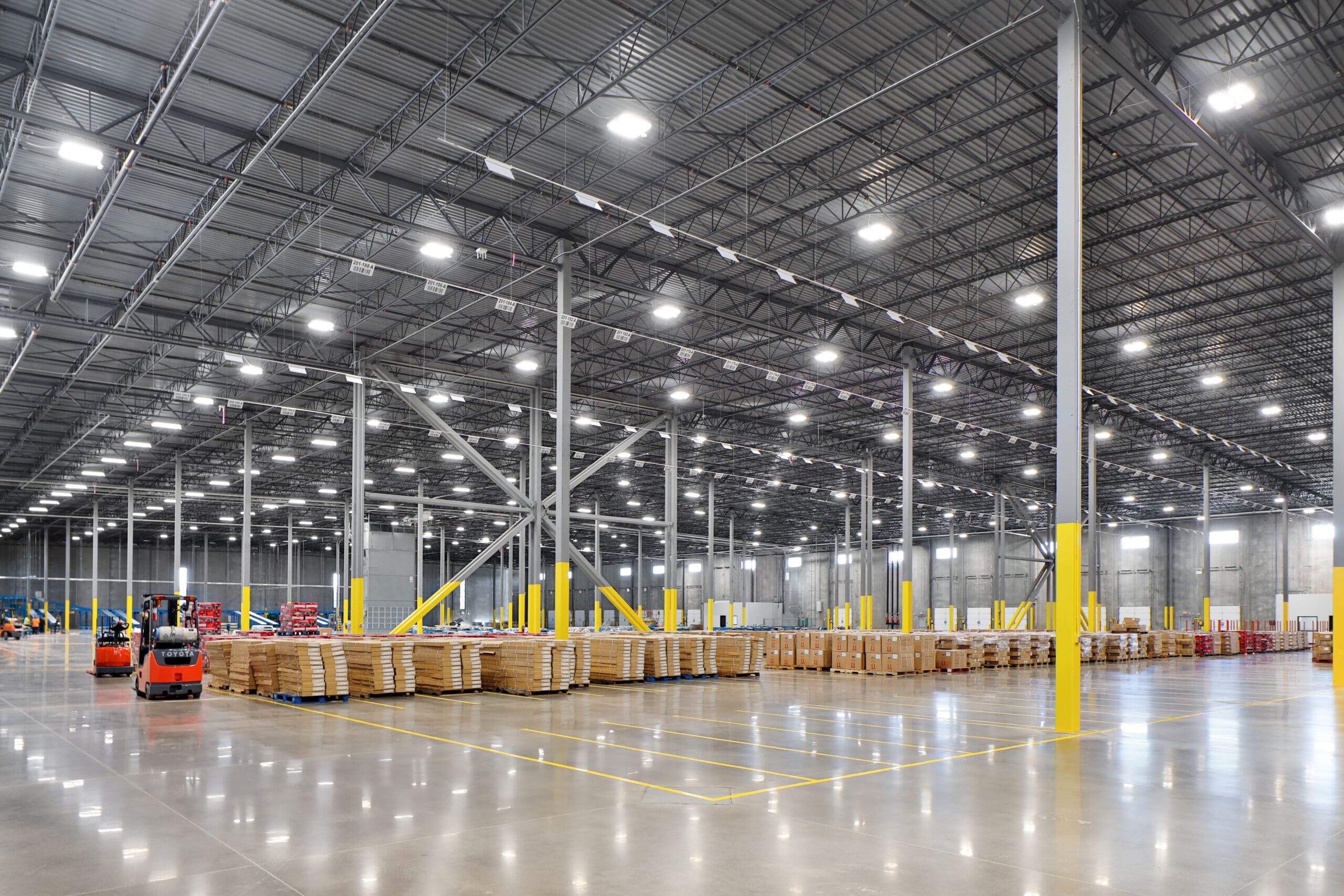 A large, brightly lit warehouse with high ceilings and yellow support pillars. Stacks of wooden pallets are neatly arranged in rows across the shiny concrete floor. Several forklifts are visible in the background.