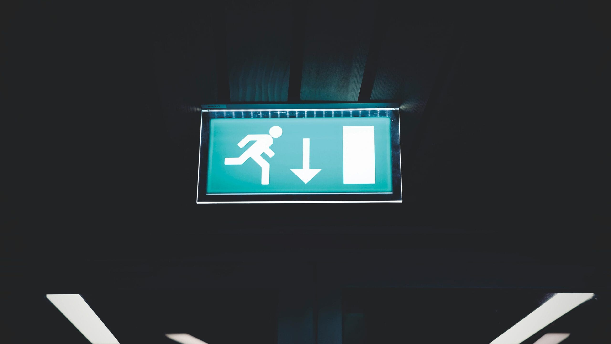 A green emergency exit sign with a running figure and a downward arrow pointing toward an open door, illuminated against a dark background.