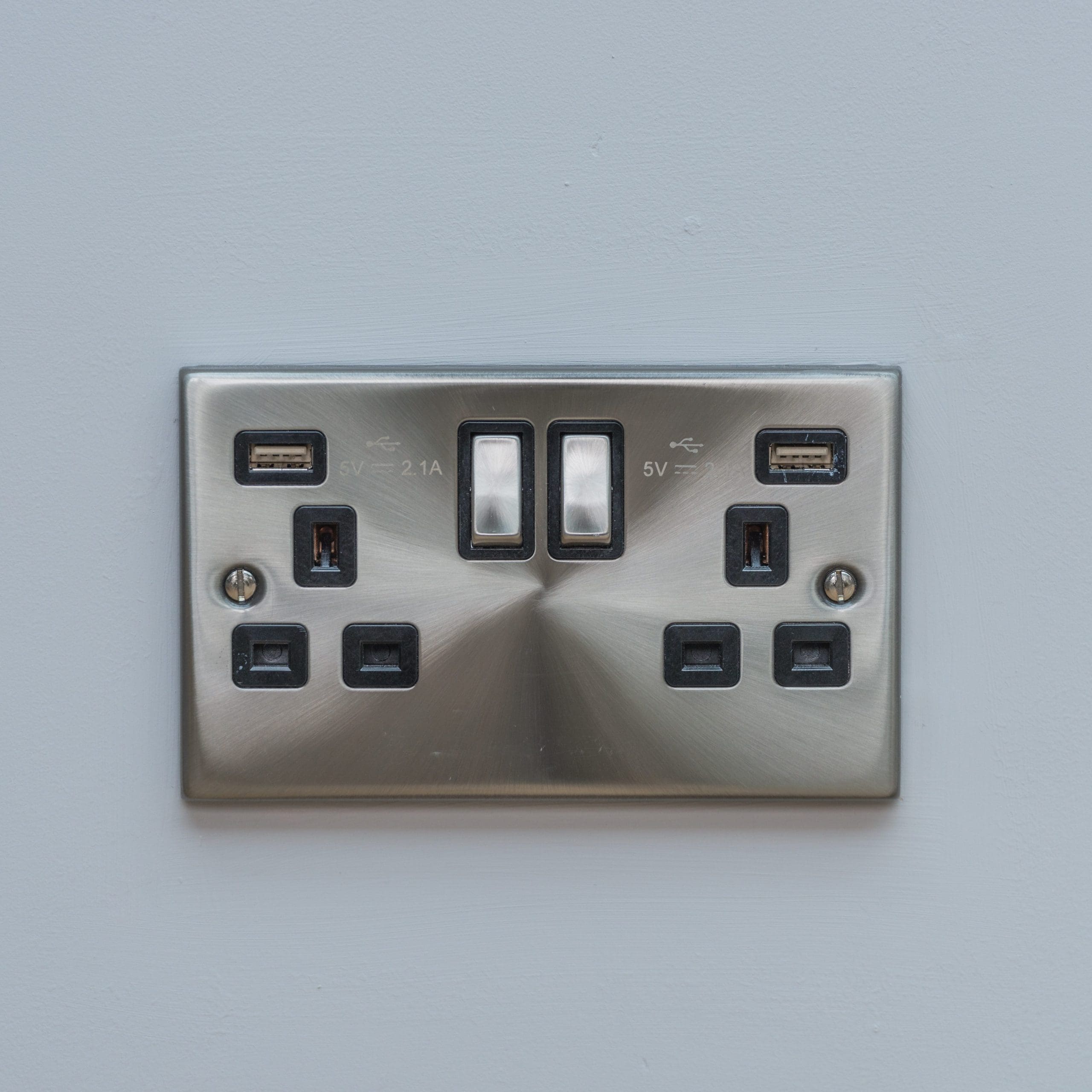 A metallic dual power socket on a white wall, featuring two USB ports above each outlet. The sockets have black switches and are labeled with charging specifications, offering 5V and 2.1A power output.