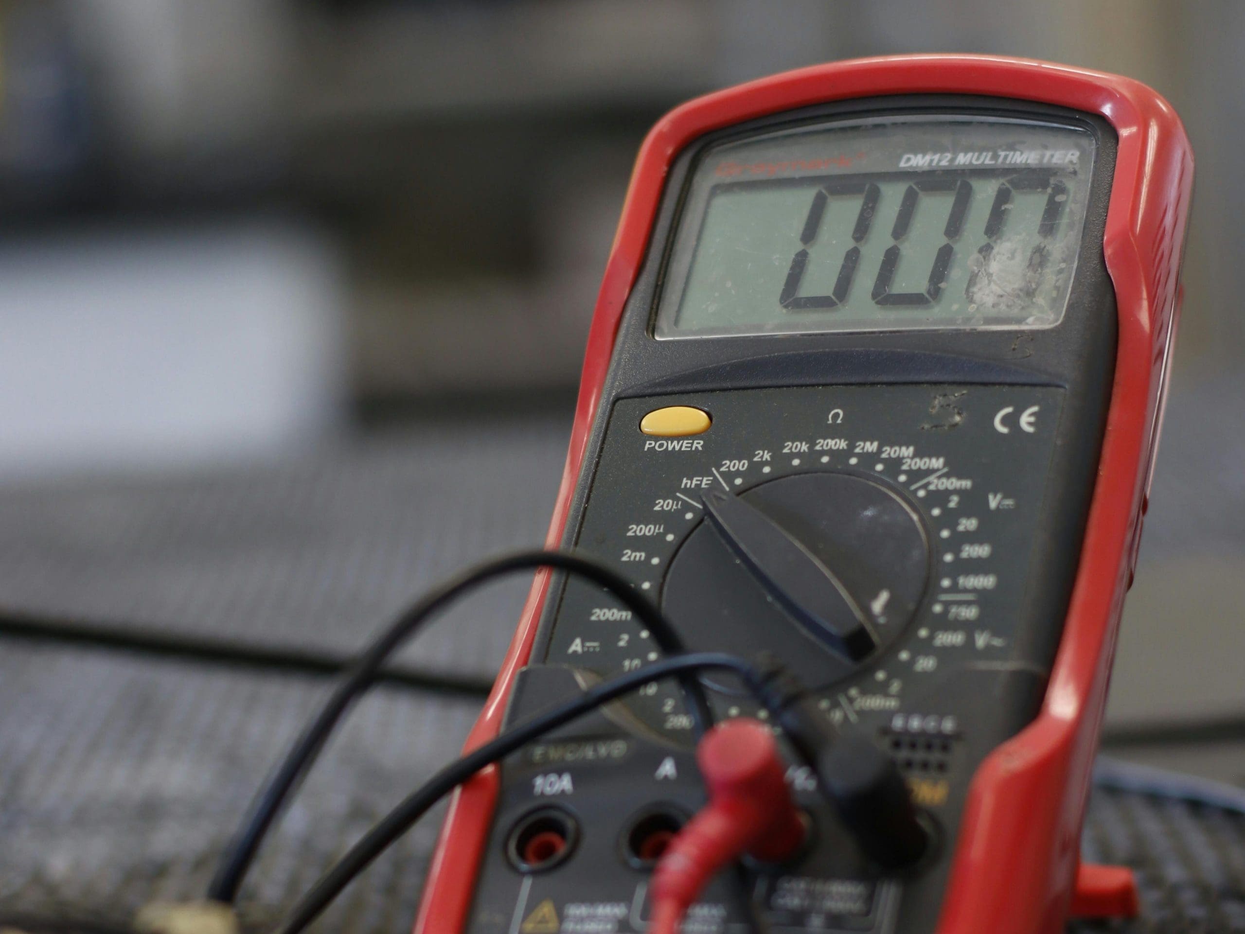A close-up of a red digital multimeter displaying "000" on the screen. The multimeter is set to measure voltage, and black and red probes are connected to it. The device is placed on a blurry, metallic surface.