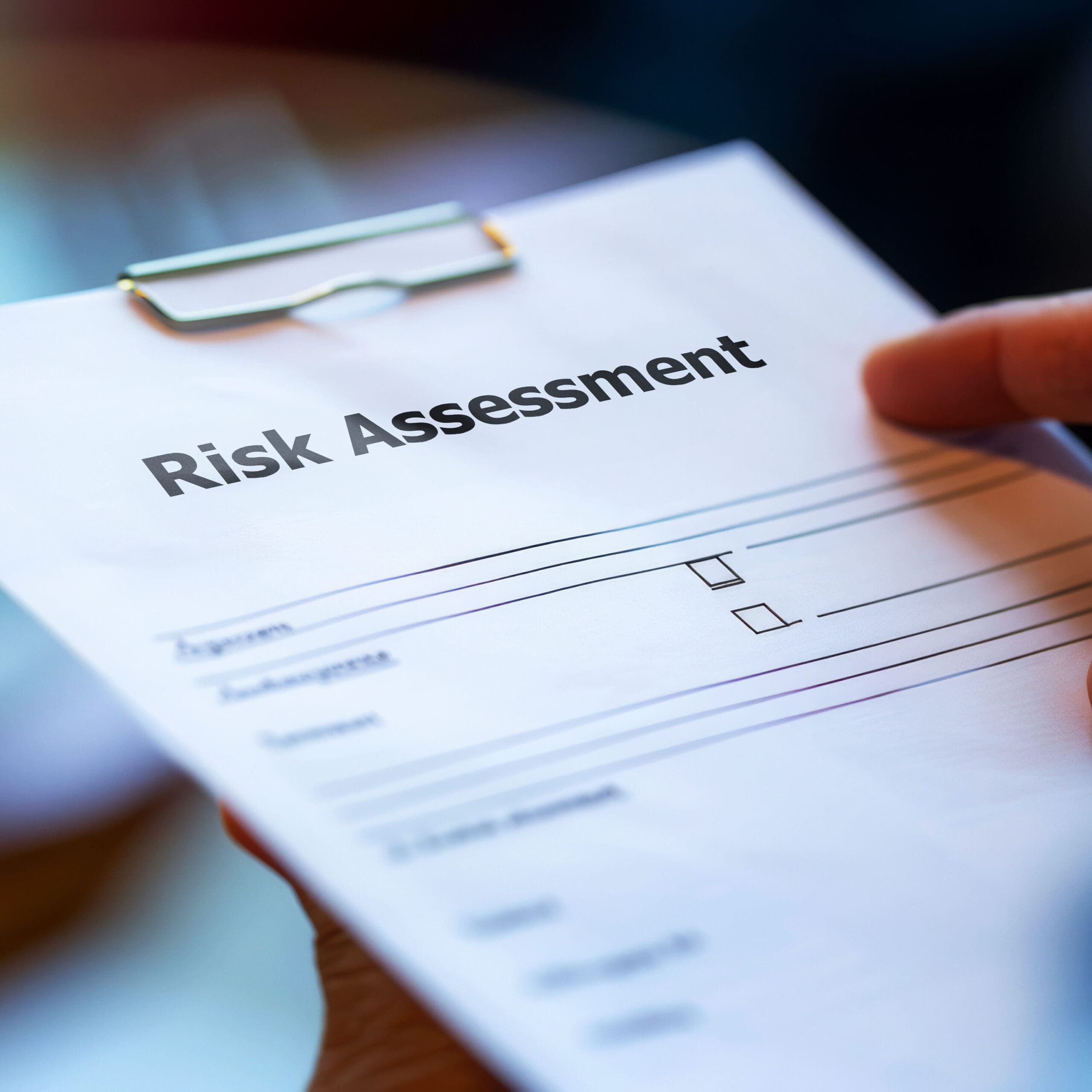 Close-up of a "Risk Assessment" document held by a person. The paper includes checkboxes and lines for input. The title is bold and prominent at the top of the page.