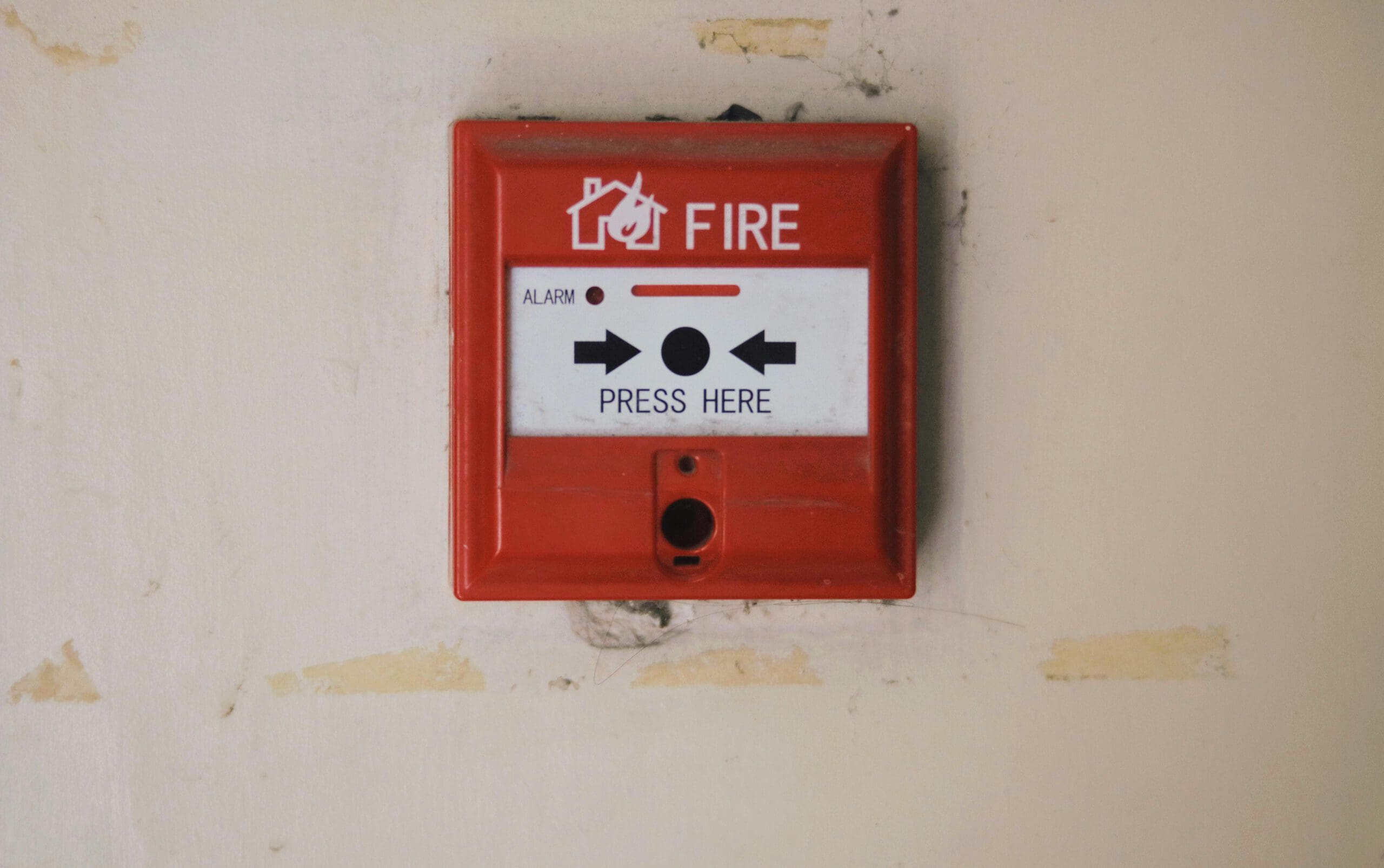 A red fire alarm box mounted on a white wall. The alarm has a label with "FIRE" and "PRESS HERE" instructions, with arrows pointing to the button. The wall shows signs of wear.