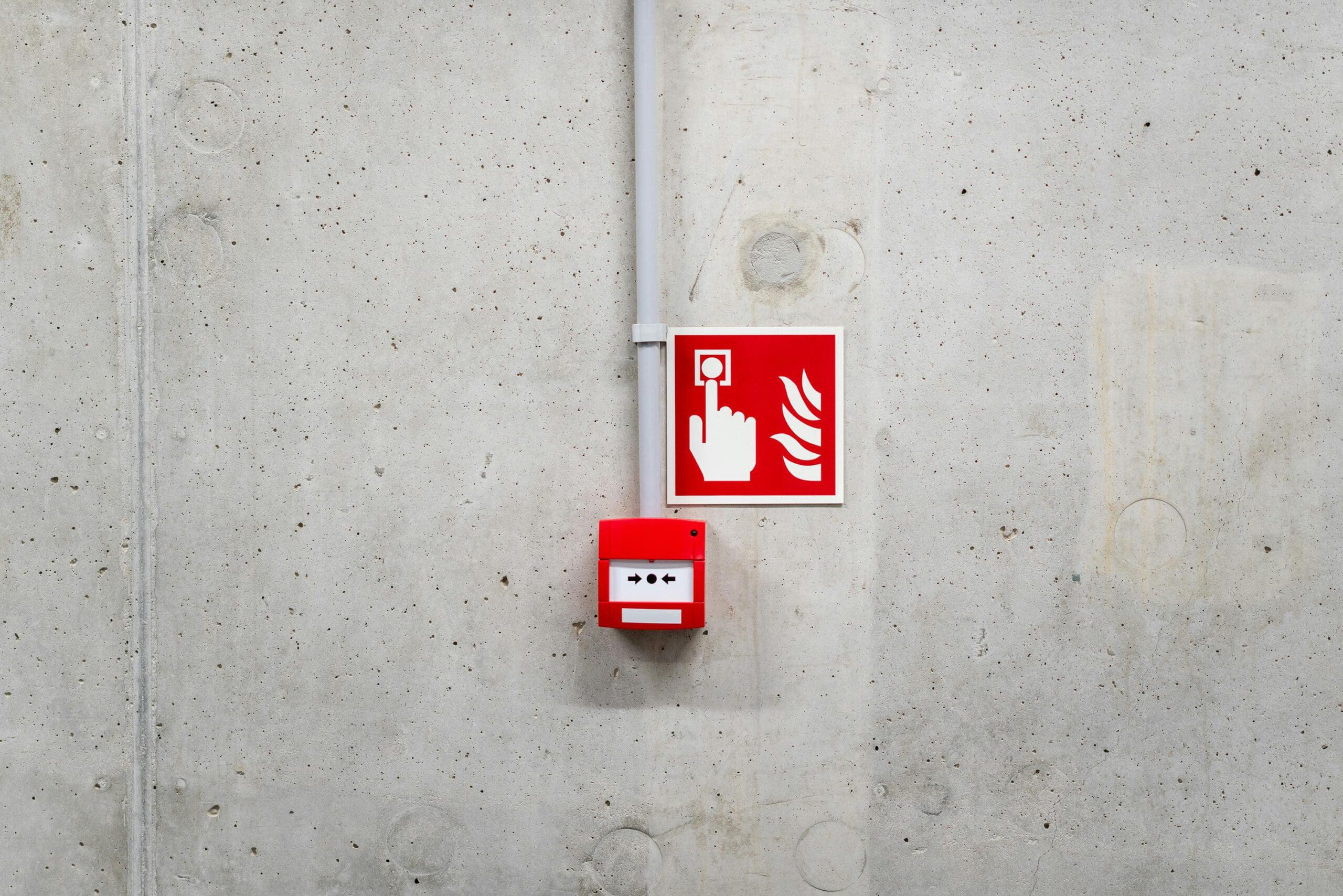 A red fire alarm button on a concrete wall, accompanied by a sign depicting a hand pressing a button with flames beside it.