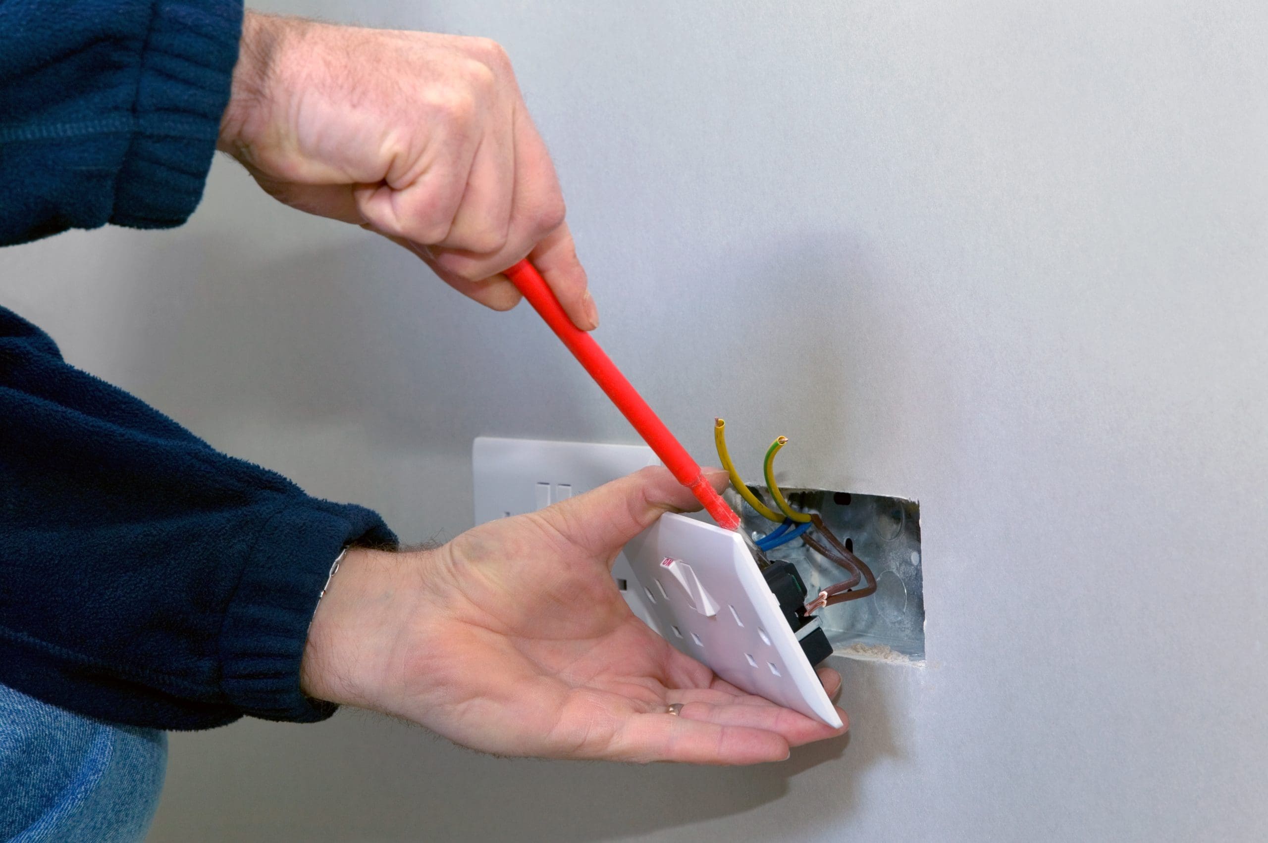A person using a screwdriver to install or remove an electrical outlet cover on a wall. Exposed wires are visible, and the person is wearing a dark long-sleeve shirt.