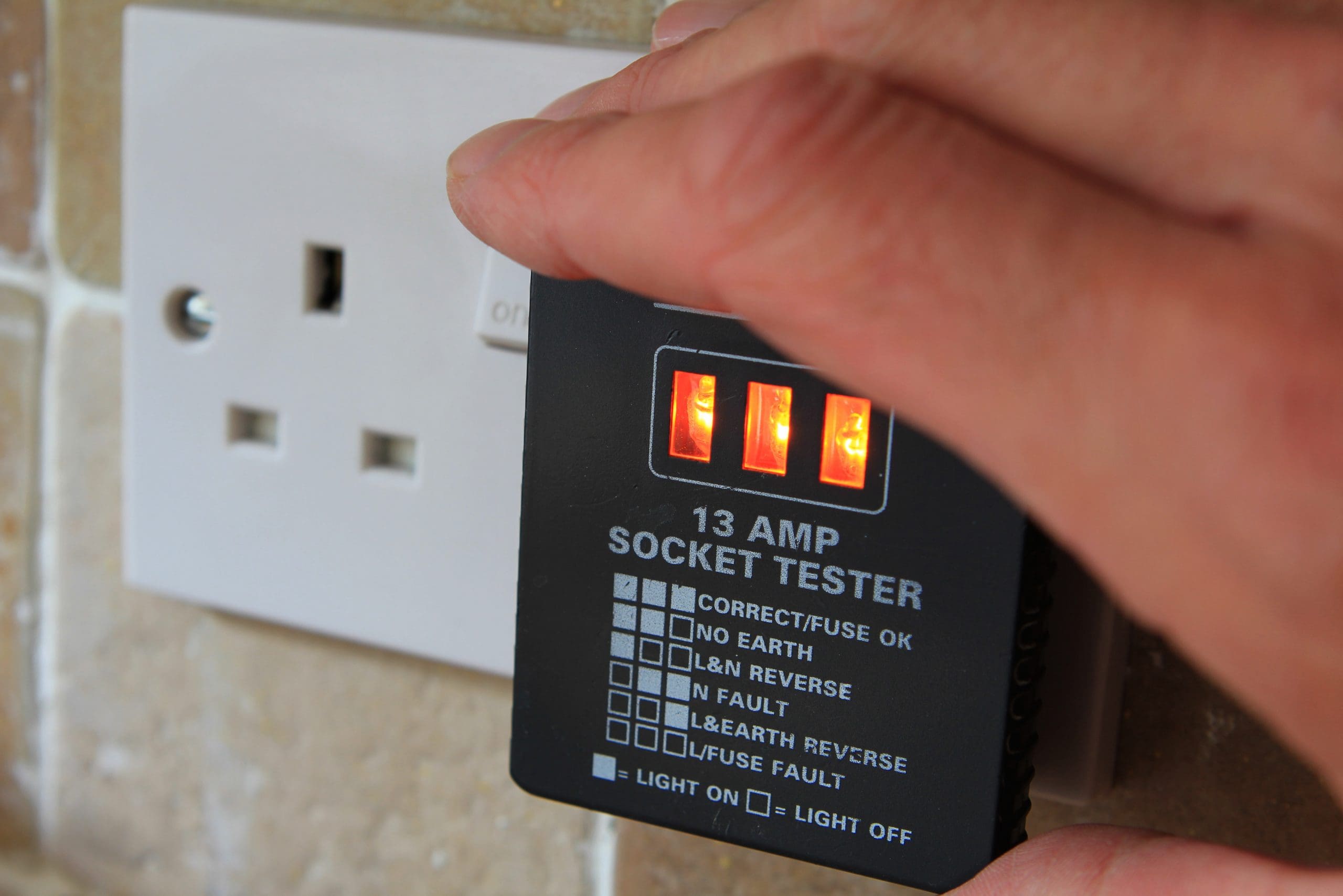 A 13 amp socket tester is being used to check a wall outlet. Three orange lights are illuminated, indicating the status of the electrical connection.