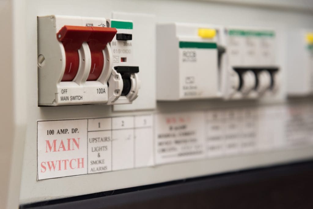 Close-up of an electrical panel featuring a red main switch labeled "100 AMP. DP. MAIN SWITCH" and several gray circuit breakers. The panel includes labels for functions like "UPSTAIRS LIGHTS & SMOKE ALARMS.