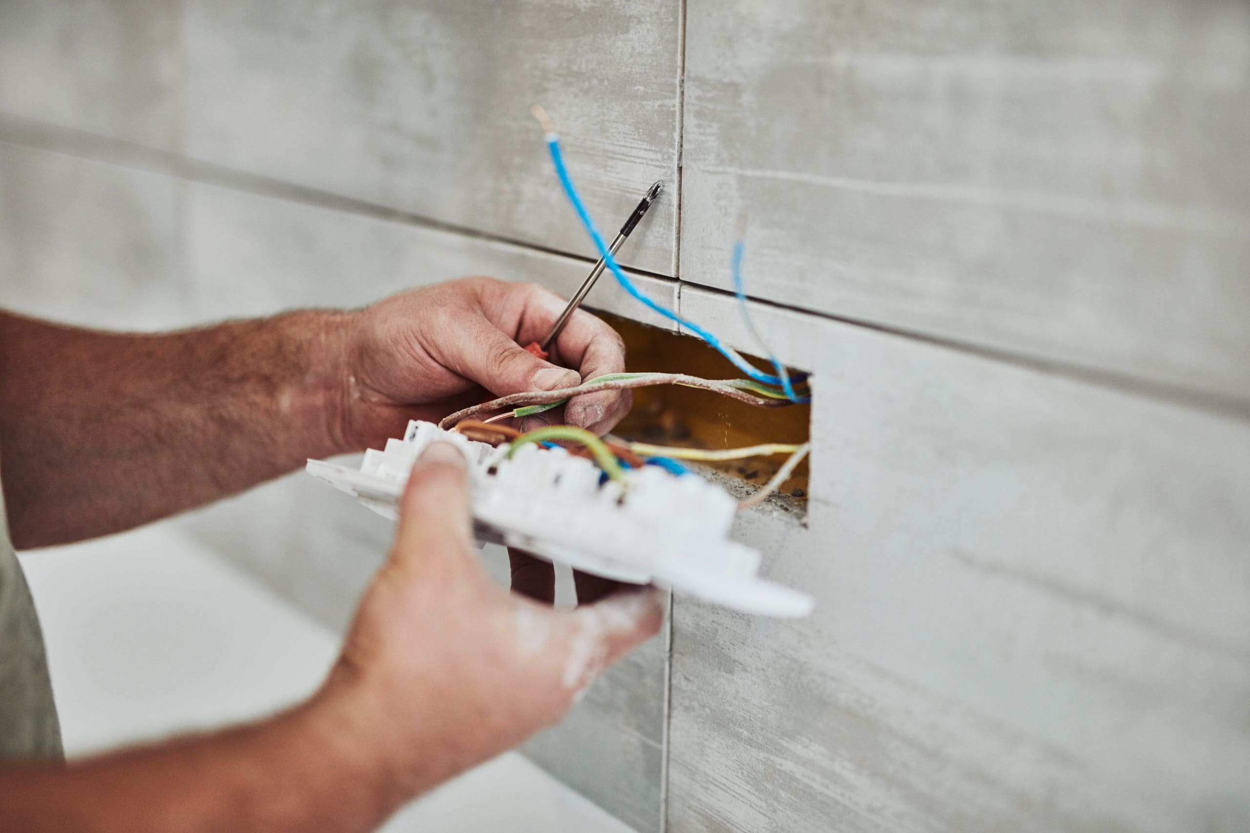 A person is installing a light switch onto a tiled wall. They are holding the switch with one hand and connecting electrical wires to it with the other hand. Several colored wires are visible within the wall opening.