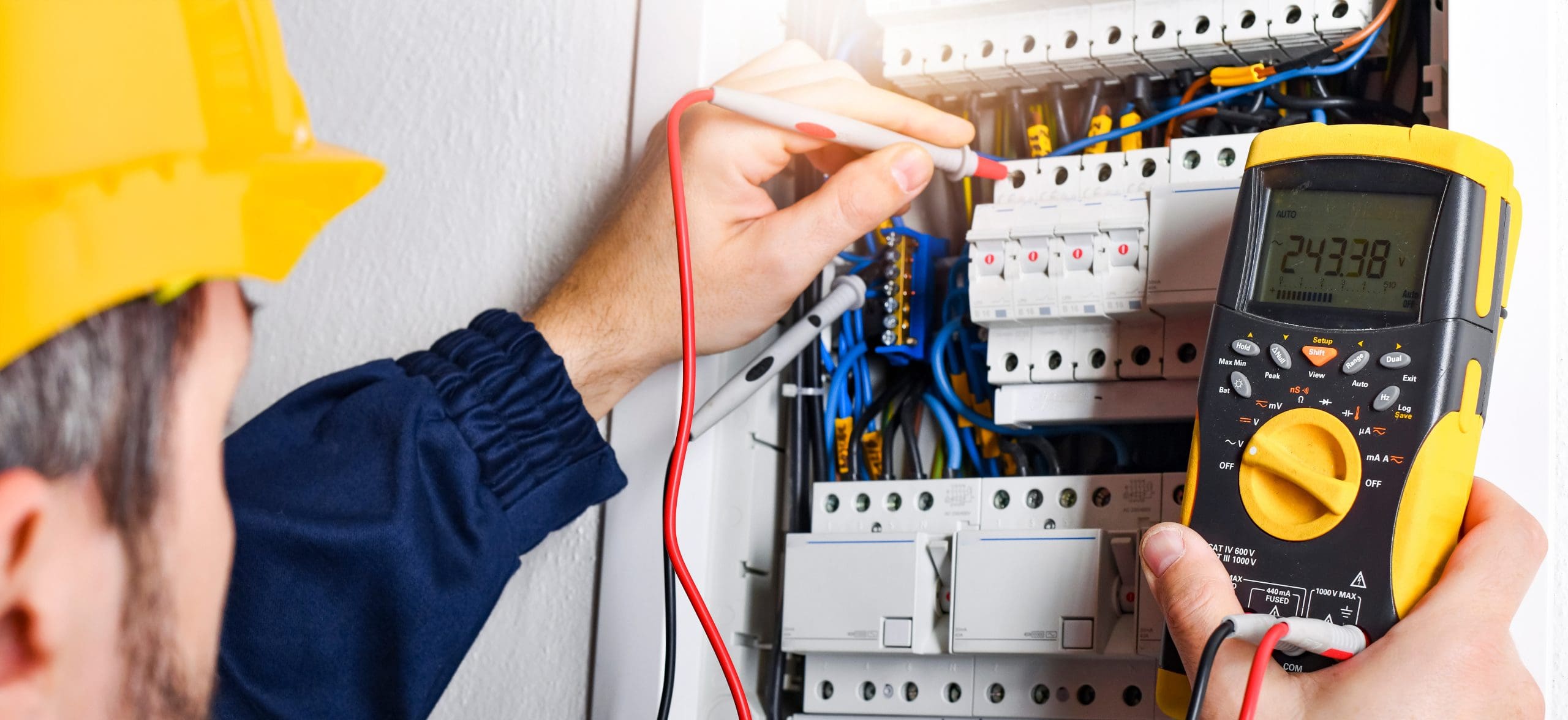 An electrician in a yellow hard hat uses a multimeter to test electrical circuits in a breaker panel filled with various colored wires. The multimeter display shows readings, indicating active measurement.