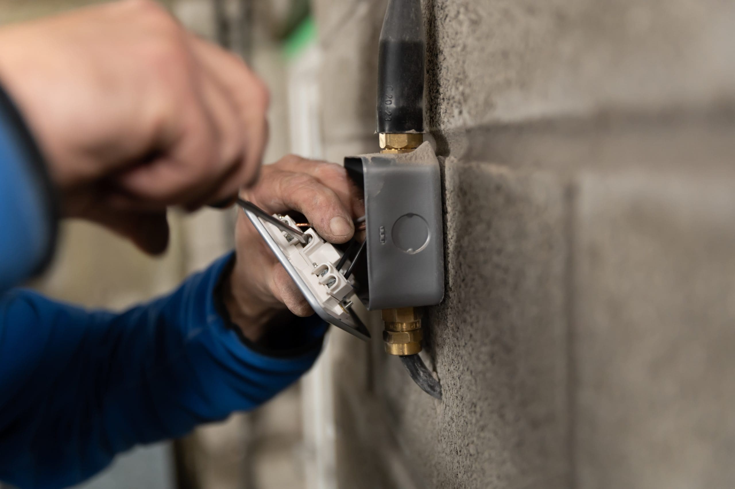 A person in a blue sleeve is installing or repairing an electrical component on a concrete wall, using a tool to adjust the wiring inside a gray junction box. Close-up view focusing on the person's hands and the component.