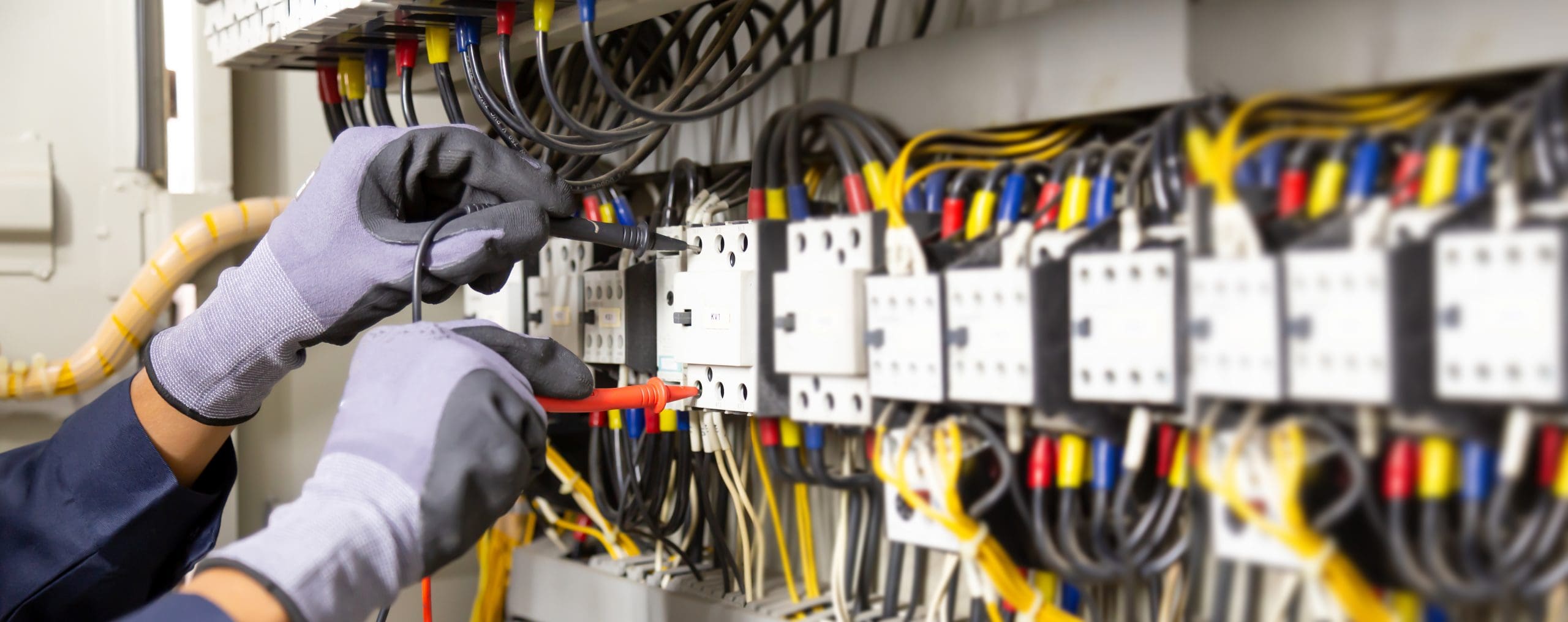 A person wearing gray gloves uses tools to adjust wiring inside an electrical panel filled with various colored wires and components.