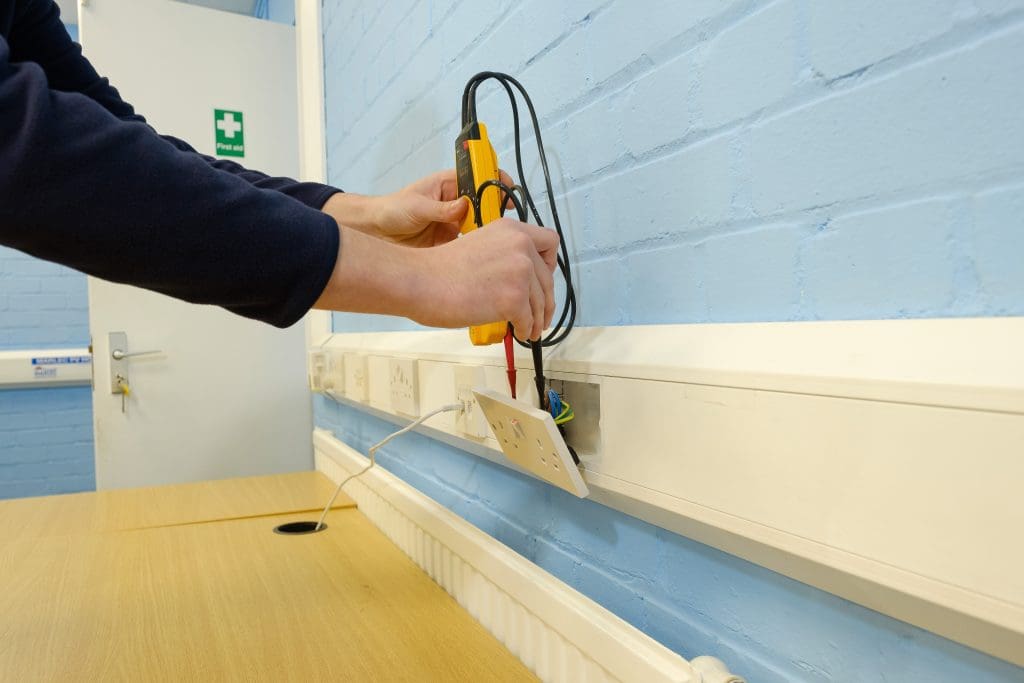 A person testing electrical wires with a multimeter in an office space. The wall is light blue, and there's a wooden desk beneath the open socket. A green first aid sign is on the background wall.